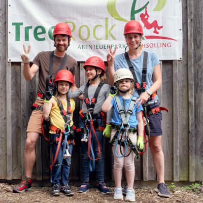 Zwei Erwachsene und drei Kinder stehen grend in Kletterausrstung vor einem groen Treerockplakat